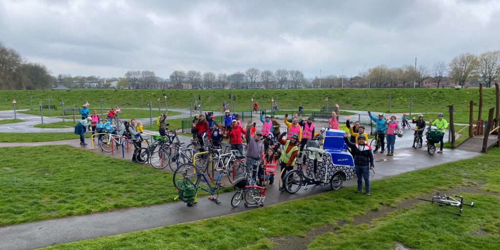 Photo of people standing and waving at the camera in a grassy park with bicycles, one of which is a large cargo bike. Some are wearing high-vis jackets. The sky is overcast and the ground is wet.