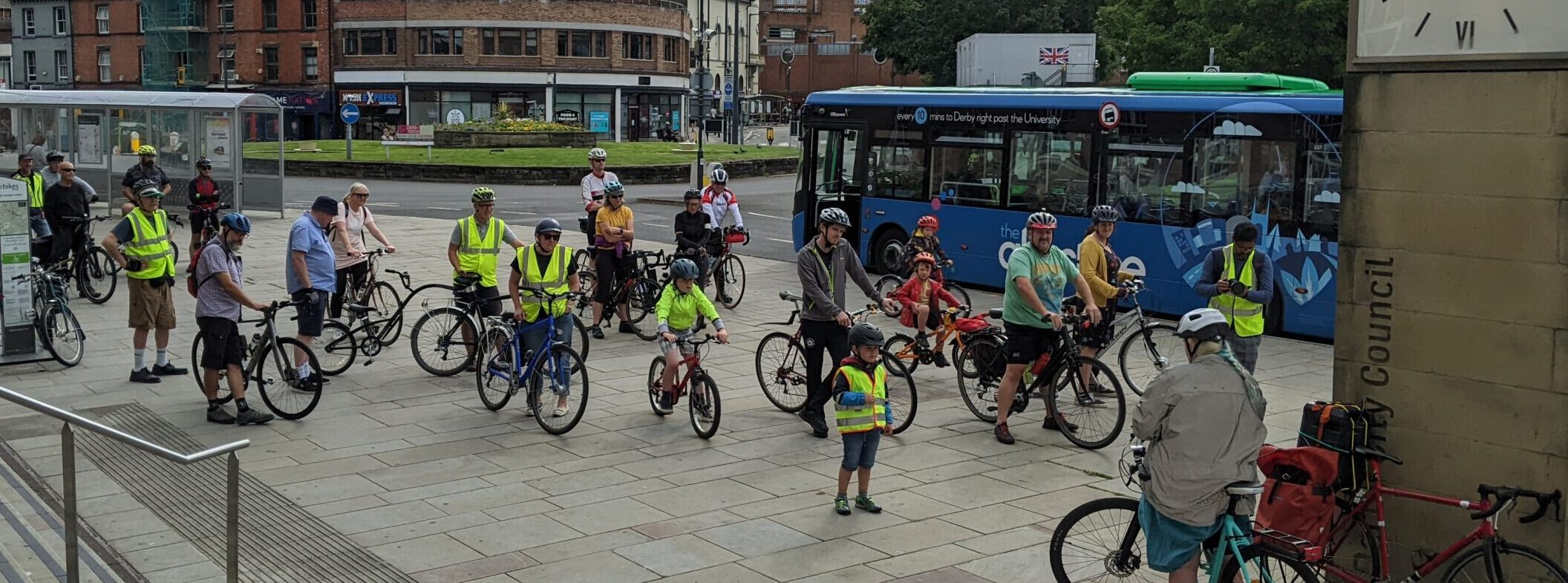 Photo: people with bicycles standing on a pavement. Behind them is a roundabout and a bus.